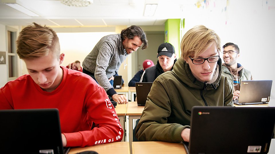 Flera elever arbetar med sina datorer i ett klassrum. En lärare instruerar en elev.