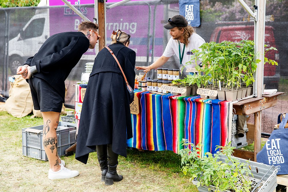 En försäljare visar sitt utbud av trädgårdsprodukter i sitt marknadsstånd för två besökare.
