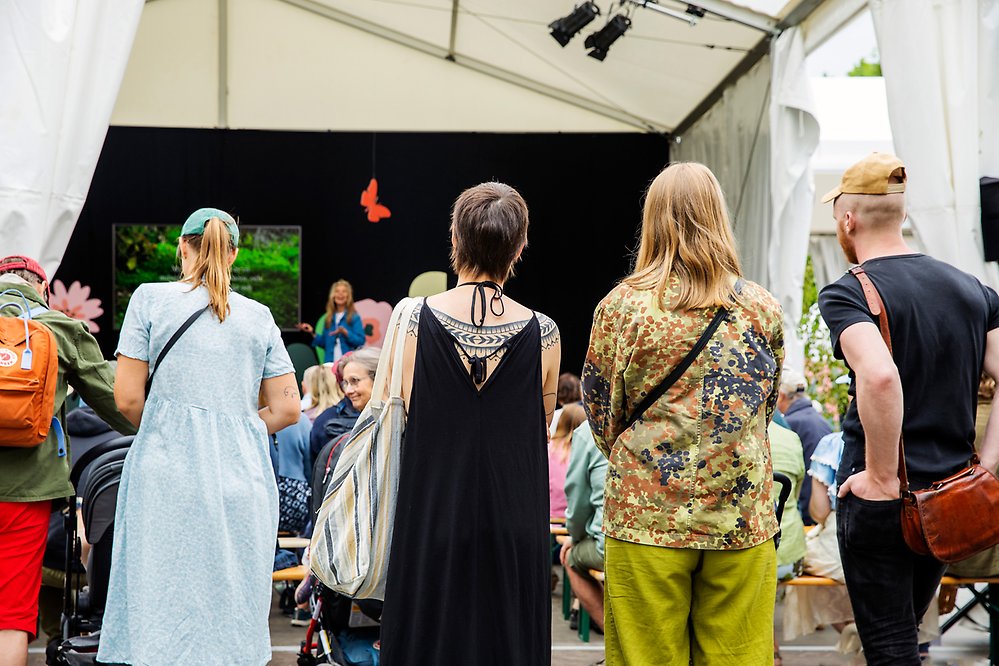 Fyra personer står och tittar på en föreläsare som pratar från en scen i ett tält inför sittande publik.