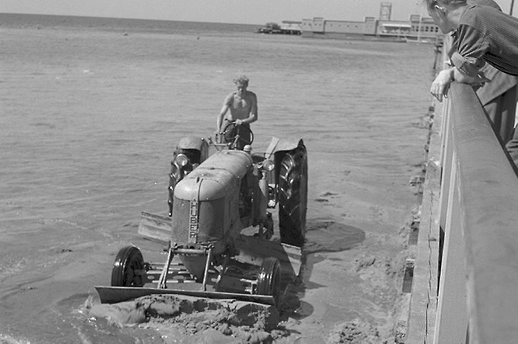 Äldre svartvitt foto. Traktor med plog kör nere i vattnet nära strand.